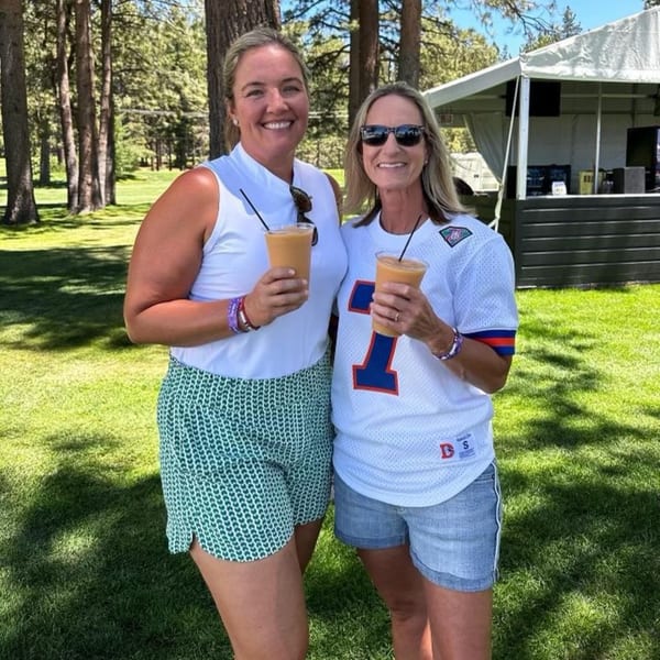 Two smiling women holding drinks outdoors, with trees and a white tent in the background.