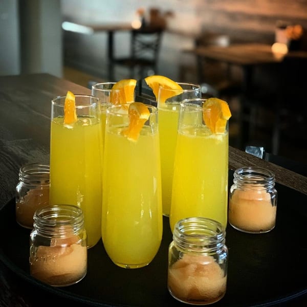 A tray of yellow mimosa cocktails garnished with orange slices and small jars of brown sugar.