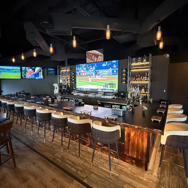 A modern bar with high-top seating, multiple large screens showing sports, and a well-stocked liquor shelf.