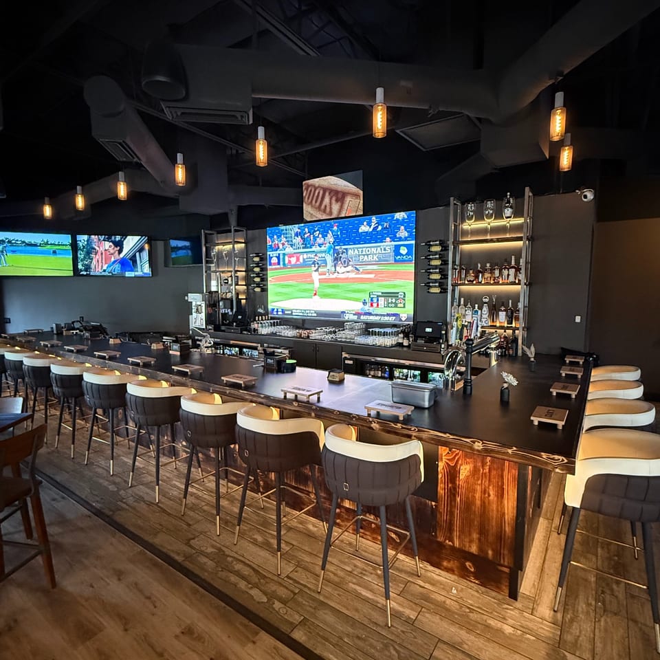 A modern bar with high-top seating, multiple large screens showing sports, and a well-stocked liquor shelf.
