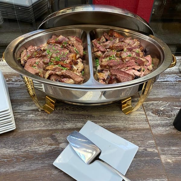 Chafing dish filled with sliced grilled meat, garnished with herbs, on a wooden table.