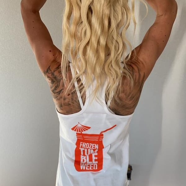A person with blonde hair and tattoos on their back wears a white tank top with a red "Frozen Tumbleweed" graphic.