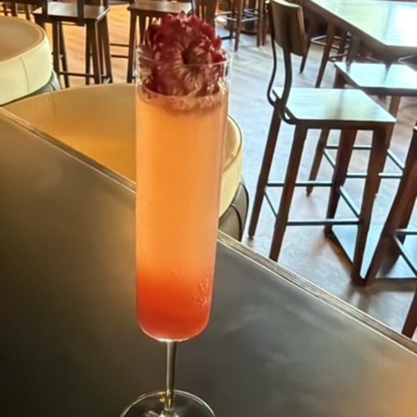 A tall, slender cocktail glass filled with an orange and red drink, garnished with red petals.