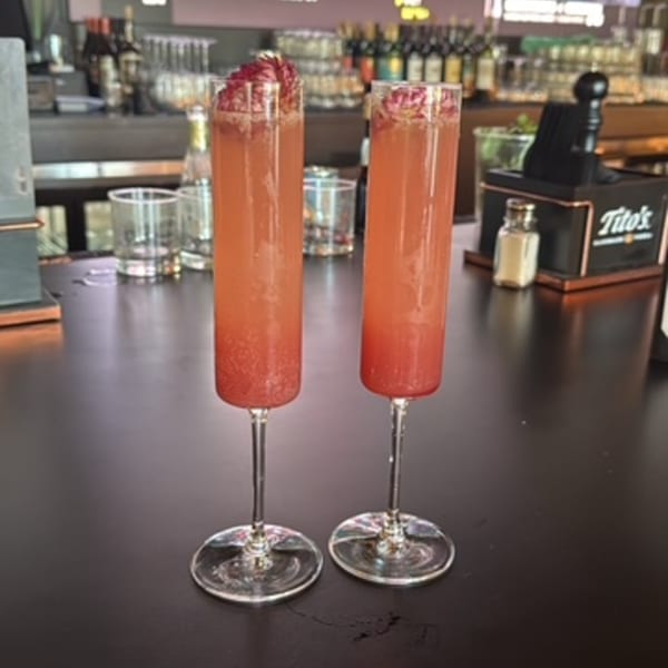 Two tall, slender glasses of pink cocktails with red garnishes on a dark bar.