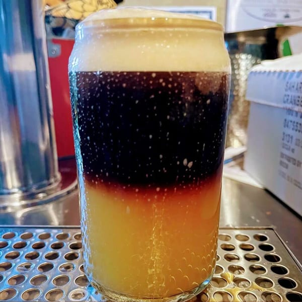 A layered drink with yellow, orange, and dark brown liquid, topped with foam, on a metal grate.