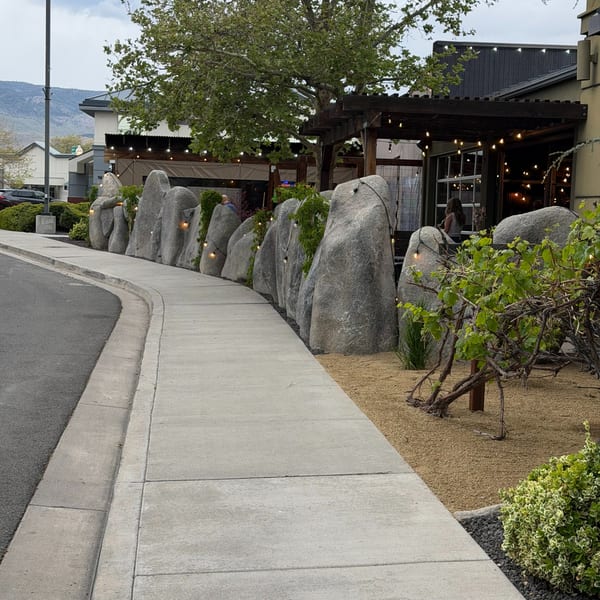 A sidewalk curves past a restaurant with a rock wall, string lights, and outdoor seating.