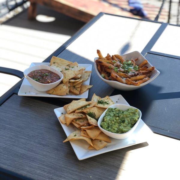 Plates of tortilla chips with salsa and guacamole, alongside a bowl of loaded fries on an outdoor table.