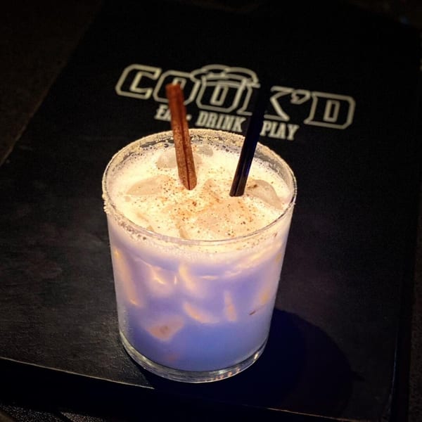 A Cook'd cocktail, light purple with ice, cinnamon stick, and black straw, on a dark surface.