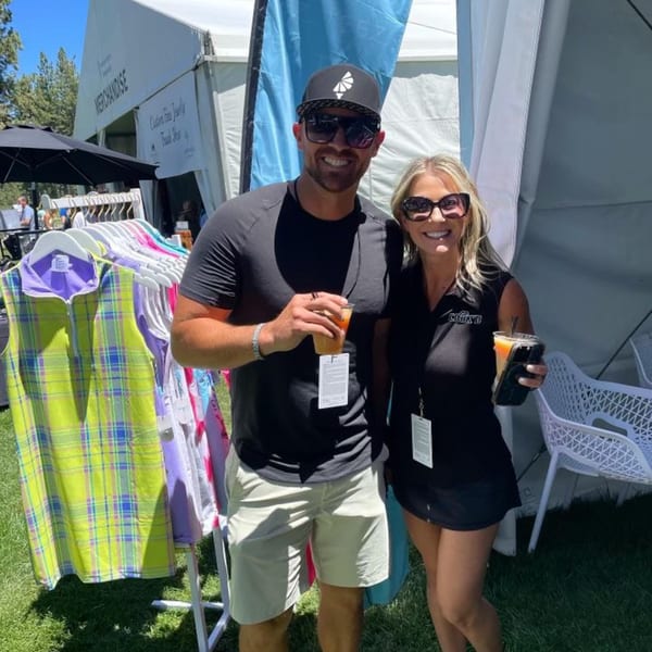 Two smiling people, a man and a woman, holding drinks next to a merchandise tent on a sunny day.