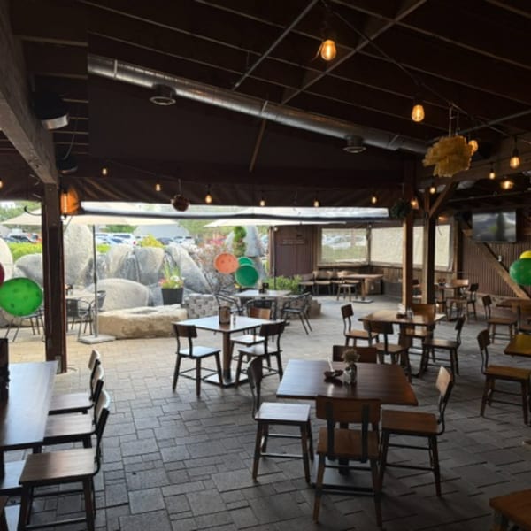 An outdoor patio dining area with wooden tables and chairs, string lights, and a rock waterfall feature.