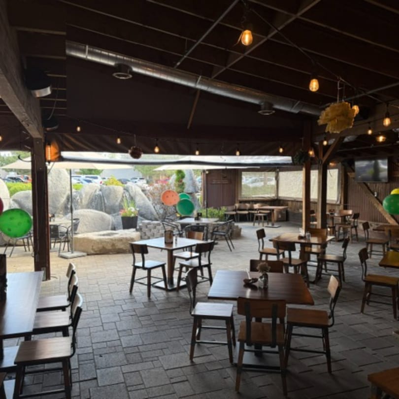 An outdoor patio dining area with wooden tables and chairs, string lights, and a rock waterfall feature.