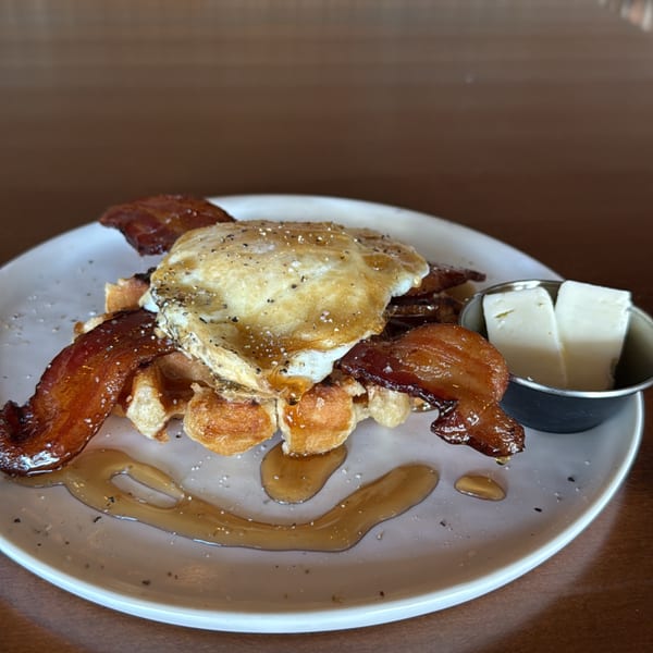 Waffle topped with crispy bacon, a fried egg, syrup, and a side of butter.