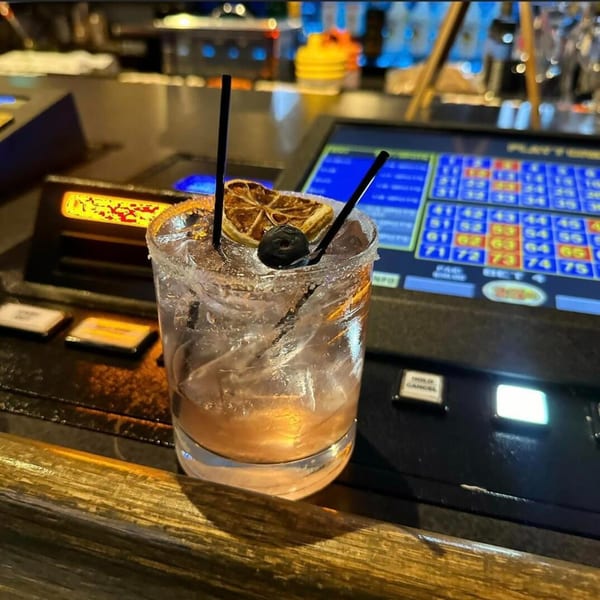 A Cook'd cocktail with a dried orange slice and blueberry, on a bar with a gaming machine.