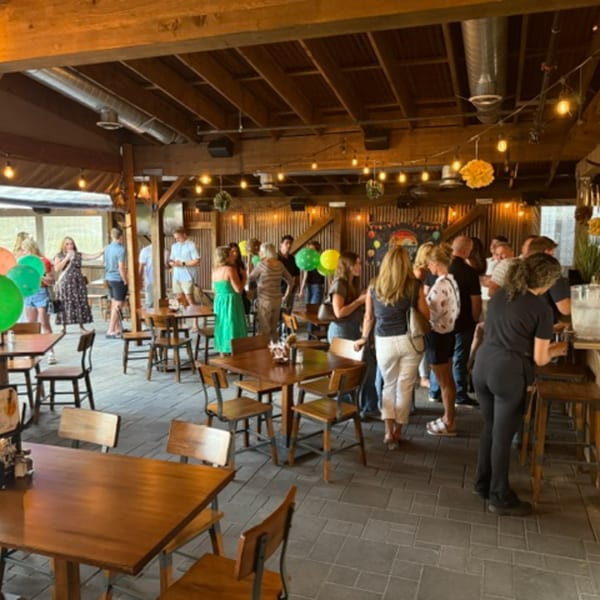 People mingling in a rustic restaurant with wooden tables, string lights, and exposed beams.