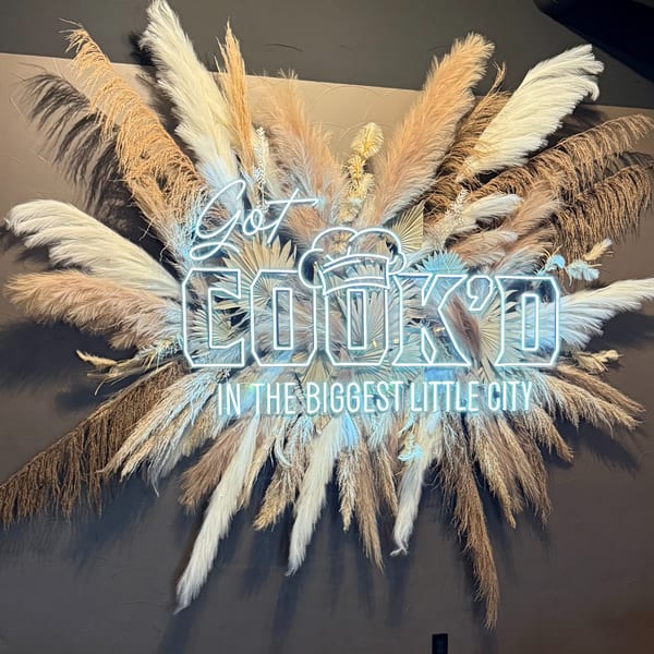 Neon sign "Got Cook'd in the Biggest Little City" surrounded by dried pampas grass.