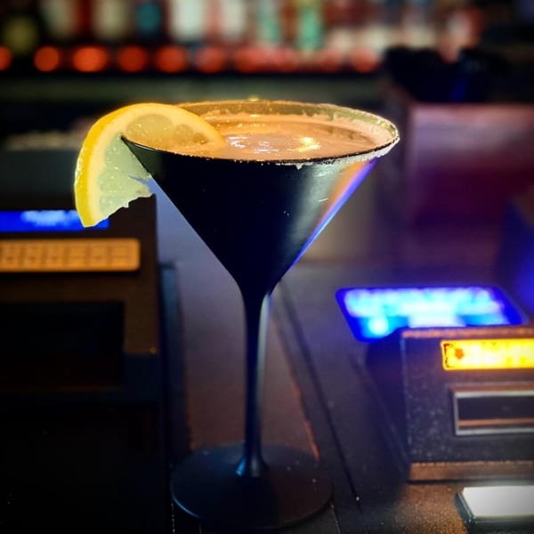 A Cook'd cocktail in a black martini glass with a lemon slice, on a bar with neon lights.