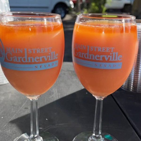 Two wine glasses filled with orange liquid, branded "Main Street Gardnerville Nevada" on a dark table.