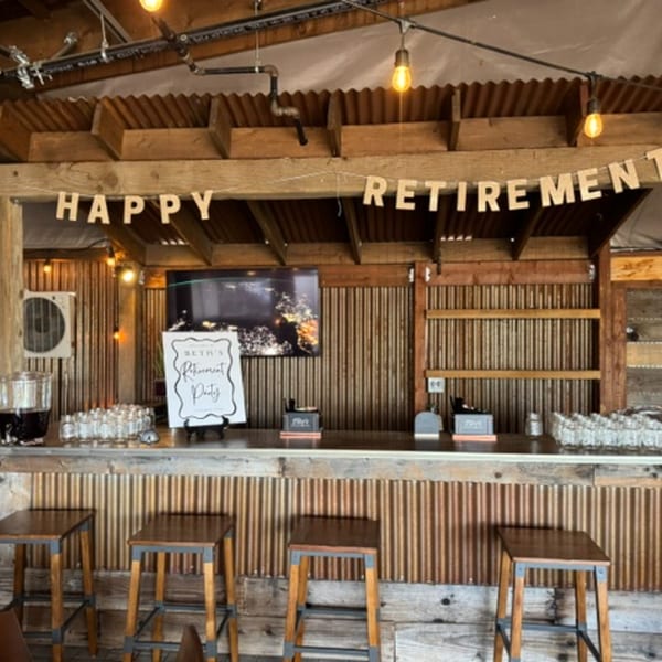 Rustic bar area with "Happy Retirement" banner, drinks, and stools, ready for a party.