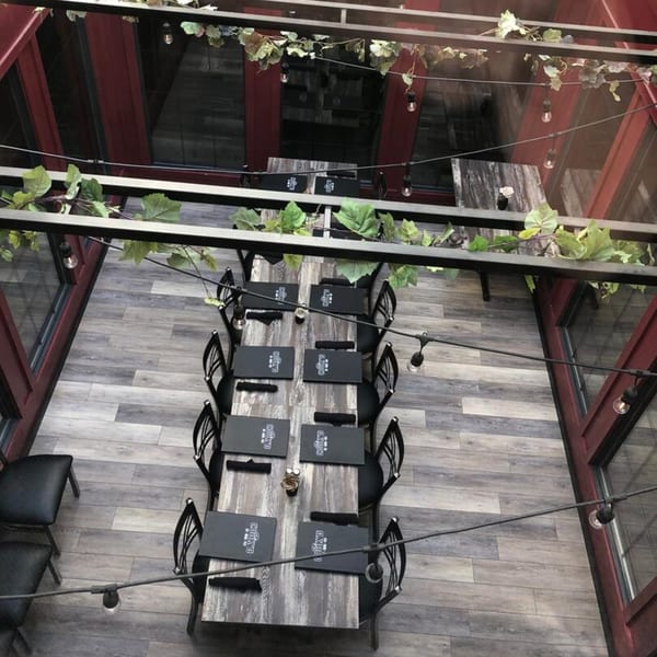 Overhead view of a long dining table set with menus, surrounded by chairs, under string lights and greenery.