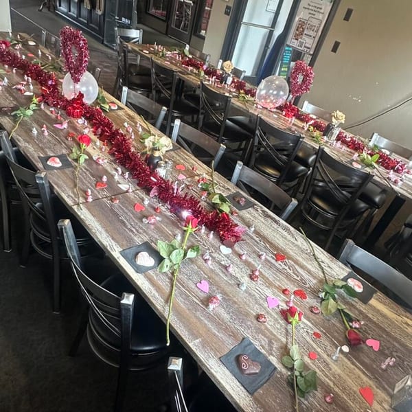 Long wooden tables decorated with red roses, heart confetti, and chocolates for a romantic setting.