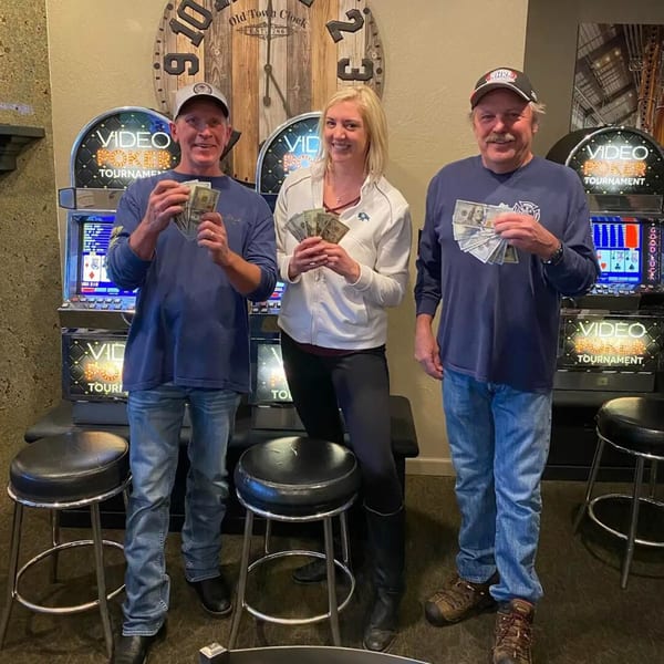 Three people holding cash prizes in front of video poker machines and a large wooden clock.