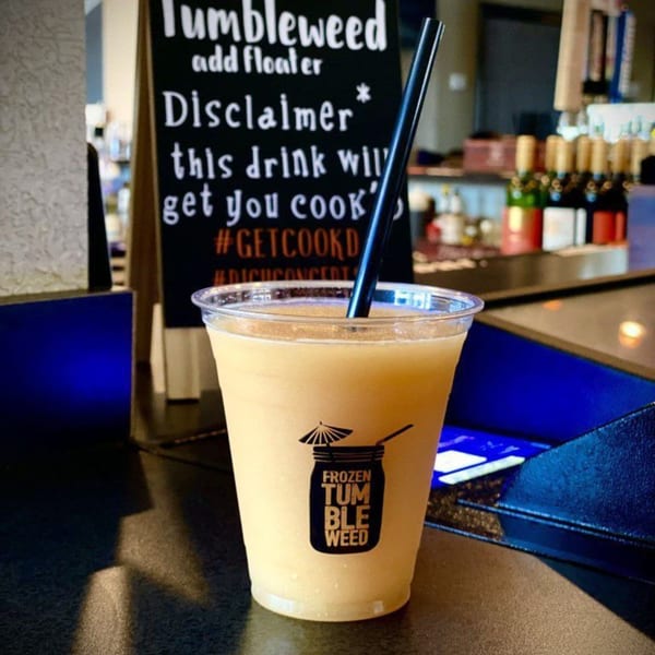 A Cook'd Frozen Tumbleweed drink with a black straw sits on a bar next to a disclaimer sign.