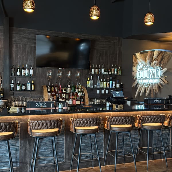A modern bar with dark wood paneling, liquor bottles, bar stools, and a neon sign.