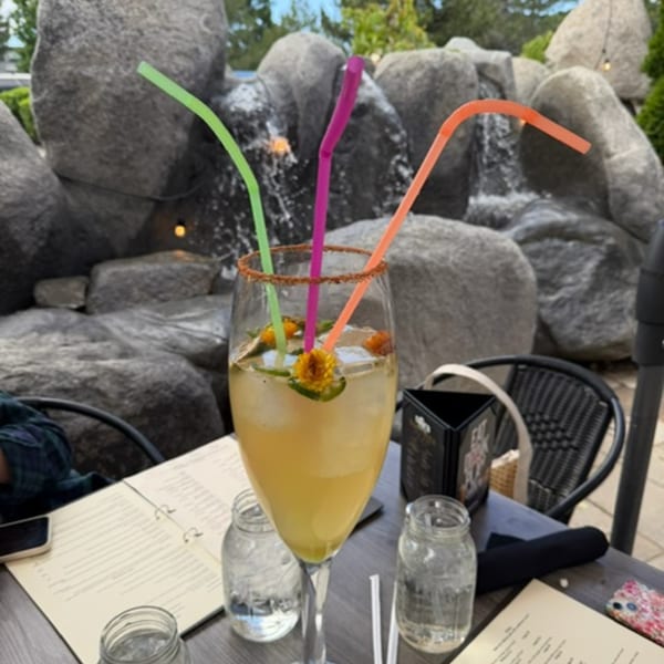 A vibrant cocktail with three colorful straws, garnished with flowers and chili rim, on an outdoor patio.