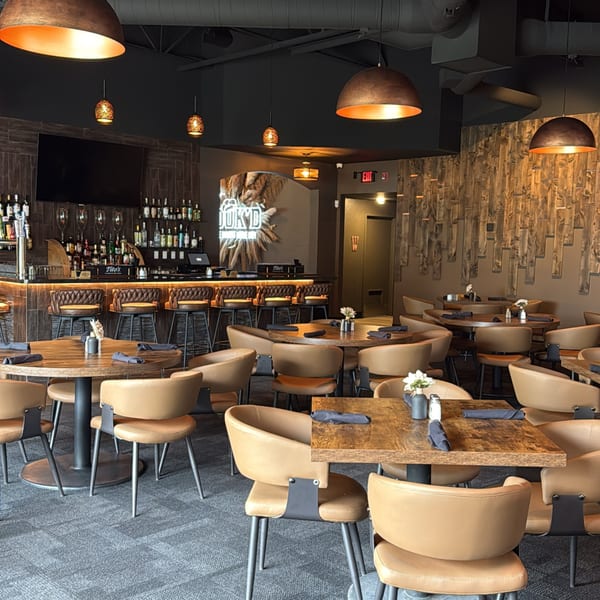 Modern restaurant interior with a full bar, wooden tables, tan chairs, and decorative lighting.