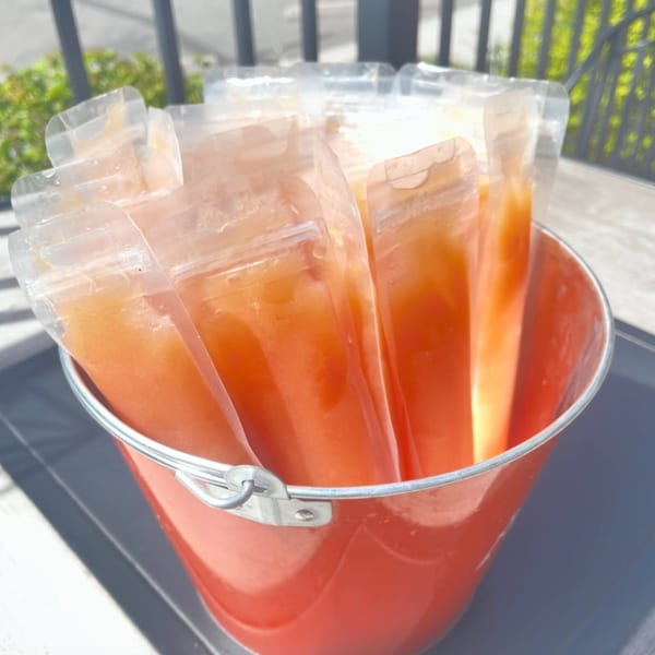 Several orange-colored frozen treats in clear pouches stand upright in a red bucket outdoors.