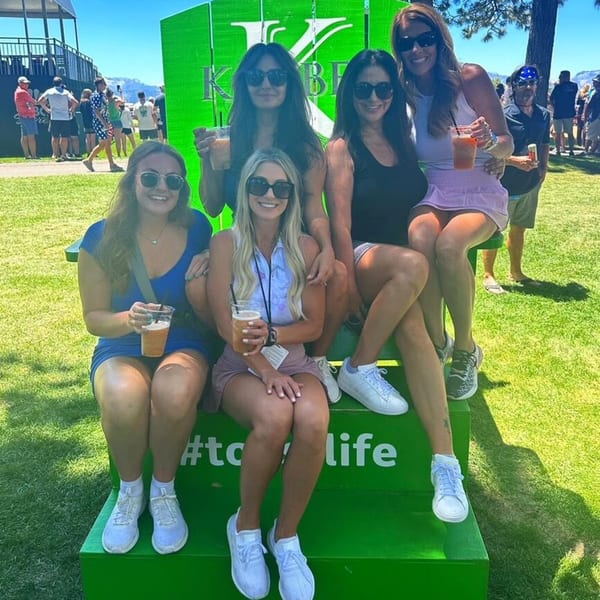 Five women in sunglasses holding drinks, posing on a green bench with a large green sign behind them.