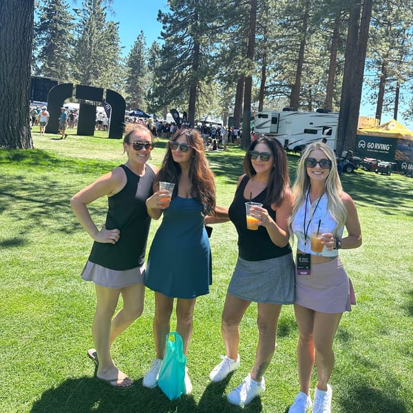 Four women in sunglasses and casual attire hold drinks on a sunny grassy field with trees and tents.