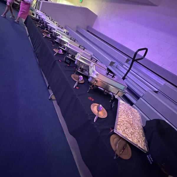 A long buffet table with chafing dishes and serving utensils, under purple lighting.