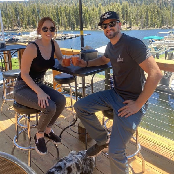 A couple smiles, holding drinks at an outdoor Cook'd restaurant table overlooking a lake with boats.