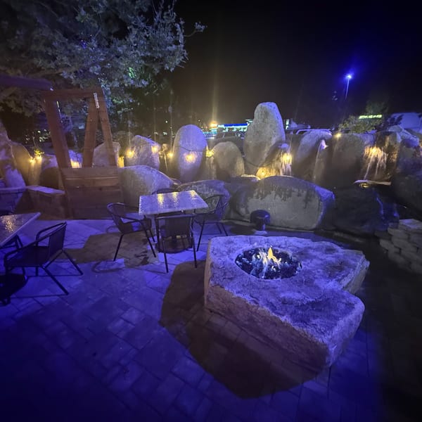 Outdoor patio at night with a fire pit, tables, chairs, and large rocks with string lights.