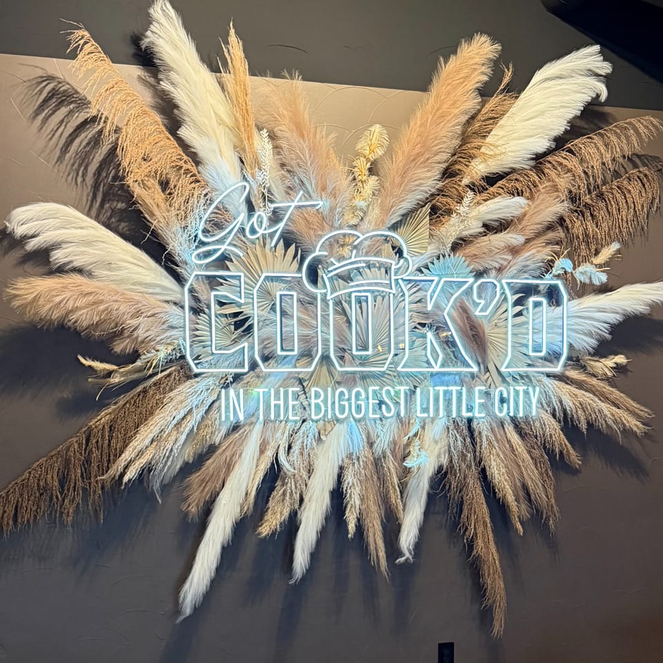 Neon sign "Got Cook'd in the Biggest Little City" surrounded by dried pampas grass on a dark wall.
