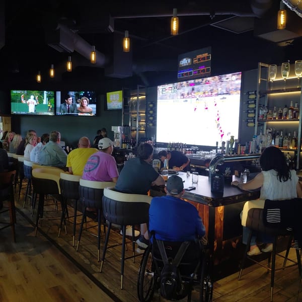 Patrons watch sports on large screens at a dark, modern bar with a bartender serving drinks.